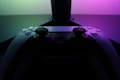 A video game controller with a sleek, modern design is positioned in front of a glowing display. The lighting casts a vibrant gradient of pink and green tones, creating a visually striking contrast against the dark backdrop.