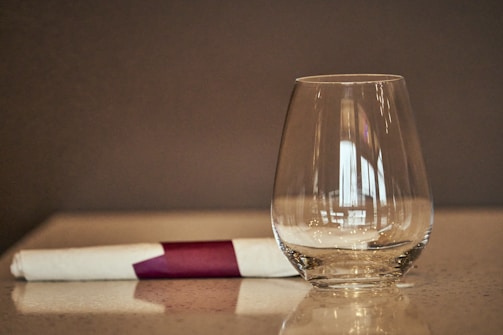 A clear water glass sits upright on a polished surface next to a neatly rolled white napkin secured with a purple band, creating a simple and elegant tabletop setting against a blurred background.