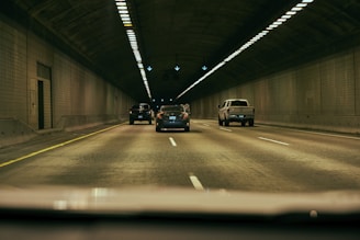 Vehicles drive through a dimly lit tunnel with tiled walls and overhead lights. The scene captures a straight road with a yellow line on the left and white dashed lines dividing lanes. Multiple vehicles are moving forward in an orderly manner.