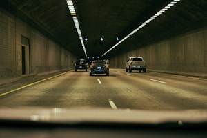 Vehicles drive through a dimly lit tunnel with tiled walls and overhead lights. The scene captures a straight road with a yellow line on the left and white dashed lines dividing lanes. Multiple vehicles are moving forward in an orderly manner.