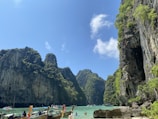A breathtaking seascape featuring towering limestone cliffs covered in lush greenery rising from turquoise waters. Several traditional wooden longboats with colorful decorations are floating on the water. A few people are visible enjoying the picturesque scenery. The sky is clear, with a few scattered clouds.