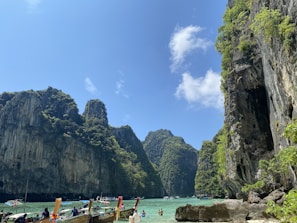 A breathtaking seascape featuring towering limestone cliffs covered in lush greenery rising from turquoise waters. Several traditional wooden longboats with colorful decorations are floating on the water. A few people are visible enjoying the picturesque scenery. The sky is clear, with a few scattered clouds.