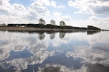 A serene landscape featuring a calm body of water reflecting the cloudy sky above. Trees line the distant shore, their silhouettes mirrored in the water, creating a tranquil and harmonious scene.