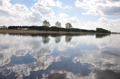 A serene landscape featuring a calm body of water reflecting the cloudy sky above. Trees line the distant shore, their silhouettes mirrored in the water, creating a tranquil and harmonious scene.