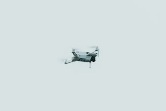 A small drone is flying against a pale, minimalistic sky. The drone appears to be lightweight, featuring four propellers and a compact design.