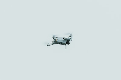 A small drone is flying against a pale, minimalistic sky. The drone appears to be lightweight, featuring four propellers and a compact design.
