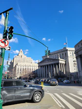 A street scene with a prominent, historic courthouse featuring large columns. The sky is clear and blue, with a few scattered clouds. Cars are parked along the street near a traffic light and no turn sign.