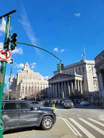 A street scene with a prominent, historic courthouse featuring large columns. The sky is clear and blue, with a few scattered clouds. Cars are parked along the street near a traffic light and no turn sign.