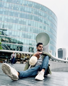 A man sits casually on a boardwalk in an urban setting, with a modern glass building in the background. He is wearing casual attire, including jeans, a green jacket, and white sneakers, while holding a beige cap in his hand. People can be seen walking in the background, contributing to the lively cityscape atmosphere.
