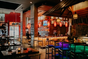 A warm and inviting dining area with a bar as the focal point, featuring a variety of hanging lanterns and decorative accents. The bar is well-stocked with bottles and has several wooden bar stools. There are tables set with glasses and small candles, creating a cozy atmosphere. The lighting is dim and ambient, adding to the relaxed mood.
