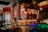 A warm and inviting dining area with a bar as the focal point, featuring a variety of hanging lanterns and decorative accents. The bar is well-stocked with bottles and has several wooden bar stools. There are tables set with glasses and small candles, creating a cozy atmosphere. The lighting is dim and ambient, adding to the relaxed mood.