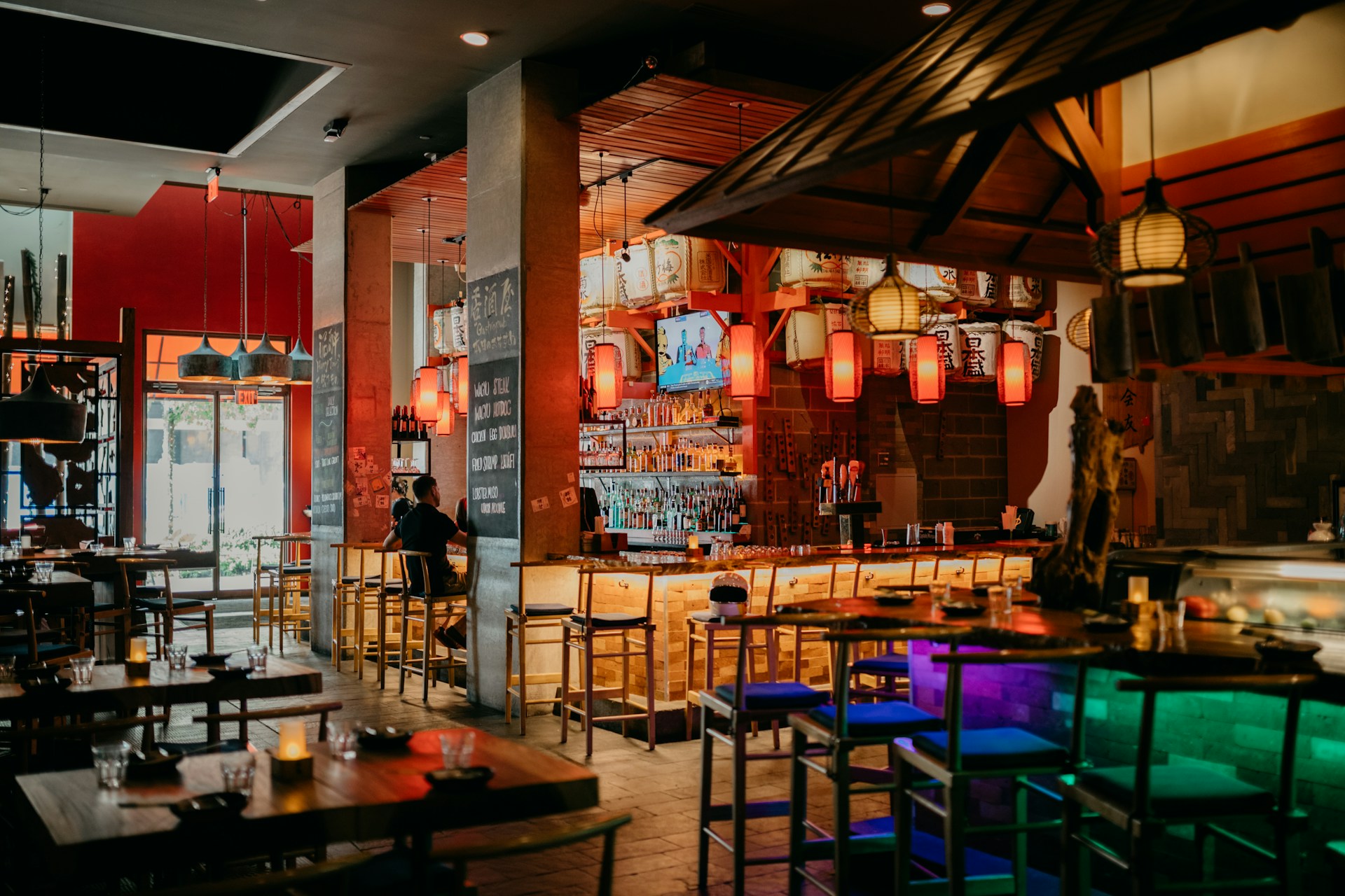 A warm and inviting dining area with a bar as the focal point, featuring a variety of hanging lanterns and decorative accents. The bar is well-stocked with bottles and has several wooden bar stools. There are tables set with glasses and small candles, creating a cozy atmosphere. The lighting is dim and ambient, adding to the relaxed mood.