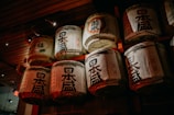 A collection of traditional Japanese sake barrels are stacked on a wooden shelf, adorned with complex kanji characters and decorative motifs. The lighting is dim, casting a warm glow over the barrels, highlighting their textured surfaces.