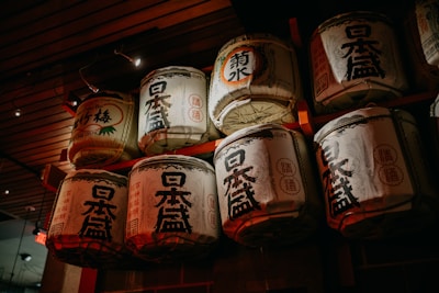 A collection of traditional Japanese sake barrels are stacked on a wooden shelf, adorned with complex kanji characters and decorative motifs. The lighting is dim, casting a warm glow over the barrels, highlighting their textured surfaces.