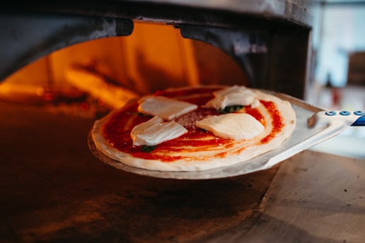 A pizza with tomato sauce and slices of mozzarella is placed on a metal peel, being inserted into a wood-fired oven with a warm glow inside.