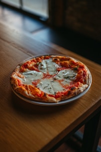 A freshly baked pizza with a crispy golden-brown crust is topped with melted cheese and visible tomato sauce. The pizza is garnished with basil leaves and is placed on a white plate atop a wooden table. Soft natural light enters from a window, creating a warm and appetizing ambiance.
