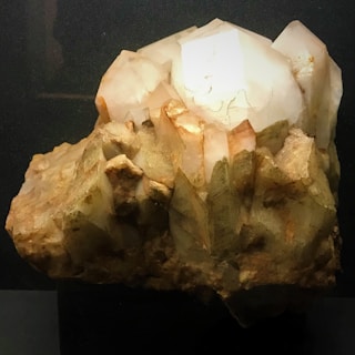A cluster of large, translucent quartz crystals with a light brown and white color palette. The crystals have various sizes and are arranged in an irregular formation. The surface is rough with visible mineral deposits, showcasing the natural beauty of the mineral.