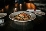 A gourmet dish is presented on a white plate, featuring a piece of grilled salmon topped with a slice of lemon. The fish is served on a bed of mixed grains and vegetables. The dimly lit, elegant setting includes a candle and a place setting with napkins and plates.