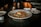 A gourmet dish is presented on a white plate, featuring a piece of grilled salmon topped with a slice of lemon. The fish is served on a bed of mixed grains and vegetables. The dimly lit, elegant setting includes a candle and a place setting with napkins and plates.