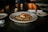 A gourmet dish is presented on a white plate, featuring a piece of grilled salmon topped with a slice of lemon. The fish is served on a bed of mixed grains and vegetables. The dimly lit, elegant setting includes a candle and a place setting with napkins and plates.