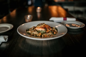 A gourmet dish is presented on a white plate, featuring a piece of grilled salmon topped with a slice of lemon. The fish is served on a bed of mixed grains and vegetables. The dimly lit, elegant setting includes a candle and a place setting with napkins and plates.