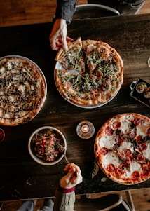 A rustic wooden table topped with a variety of pizzas and a bowl of salad. One hand is reaching to pick a slice from a pizza covered with arugula and prosciutto, while another hand is holding a fork over a quinoa salad. In the middle is a lit candle, adding a warm ambiance.