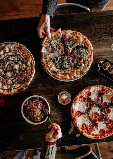 A rustic wooden table topped with a variety of pizzas and a bowl of salad. One hand is reaching to pick a slice from a pizza covered with arugula and prosciutto, while another hand is holding a fork over a quinoa salad. In the middle is a lit candle, adding a warm ambiance.
