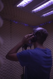 A person is standing in a soundproof recording booth wearing headphones and speaking into a microphone. The lighting in the room is dim with a purple hue coming from the overhead lights. The walls are covered in acoustic foam panels.