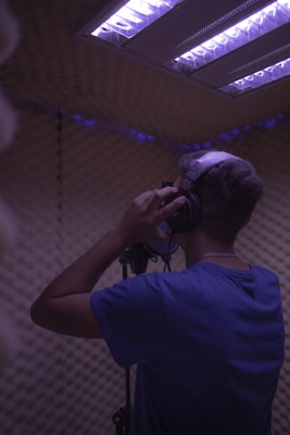 A person is standing in a soundproof recording booth wearing headphones and speaking into a microphone. The lighting in the room is dim with a purple hue coming from the overhead lights. The walls are covered in acoustic foam panels.