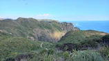 A panoramic view of lush green hills meeting turquoise sea, with a winding trail.