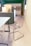 A modern office space featuring a clear, sleek acrylic chair with a chrome frame, positioned near a white table. On the opposite side, a cabinet with a vase containing a plant and a few decorative items is visible. The background includes built-in cabinets with a minimalist design.