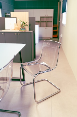 A modern office space featuring a clear, sleek acrylic chair with a chrome frame, positioned near a white table. On the opposite side, a cabinet with a vase containing a plant and a few decorative items is visible. The background includes built-in cabinets with a minimalist design.