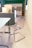 A modern office space featuring a clear, sleek acrylic chair with a chrome frame, positioned near a white table. On the opposite side, a cabinet with a vase containing a plant and a few decorative items is visible. The background includes built-in cabinets with a minimalist design.
