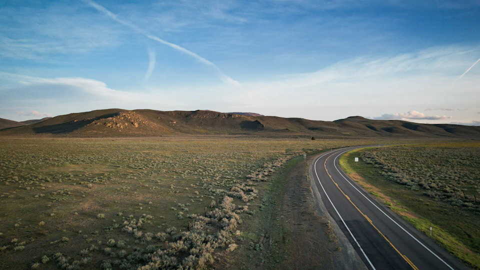 The image presents a serene desert landscape with an open road cutting through it, flanked by two hills on either side. The road is marked by a white dividing line and stretches out into the distance, disappearing beyond the horizon. On both sides of the road are patches of grass and shrubs, adding a touch of green to the otherwise barren terrain.

The sky above is a clear blue with wispy clouds scattered across it, casting long shadows on the ground below. The image is taken from an aerial perspective, providing a bird's eye view of this expansive desert landscape.