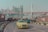 A yellow taxi drives along a road with a city skyline and a red suspension bridge in the background. Tall modern buildings are visible across the river. There are advertisements on the road barriers and vehicles on the road.