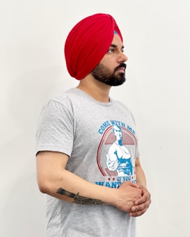 A person with a red turban and a beard is wearing a gray T-shirt featuring a graphic and text. The person is standing against a plain white background, with their hands clasped in front.