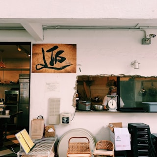 A rustic bakery scene featuring a white wall with a cutout window looking into a kitchen. A person is visible preparing food next to a scale. Various items like baskets, boxes, and a digital scale are arranged below the window. A large sign with artistic writing is mounted on the wall. The area conveys a quaint, homely atmosphere with warm tones.