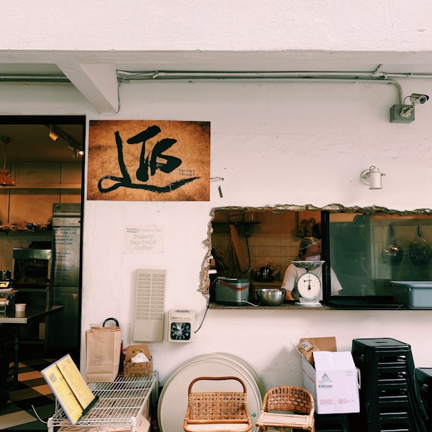 A rustic bakery scene featuring a white wall with a cutout window looking into a kitchen. A person is visible preparing food next to a scale. Various items like baskets, boxes, and a digital scale are arranged below the window. A large sign with artistic writing is mounted on the wall. The area conveys a quaint, homely atmosphere with warm tones.