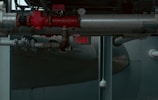 A network of industrial pipes with metallic silver and red valves, gauges, and joints runs through a dimly lit space. An illuminated exit sign is visible in the background, casting a faint red glow. The environment appears to be part of a factory or an industrial facility.