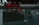 A network of industrial pipes with metallic silver and red valves, gauges, and joints runs through a dimly lit space. An illuminated exit sign is visible in the background, casting a faint red glow. The environment appears to be part of a factory or an industrial facility.