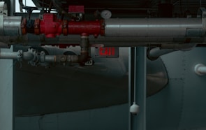 A network of industrial pipes with metallic silver and red valves, gauges, and joints runs through a dimly lit space. An illuminated exit sign is visible in the background, casting a faint red glow. The environment appears to be part of a factory or an industrial facility.