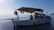 A small boat is floating on calm water, with a clear blue sky above. The boat has a canopy providing shade to a person seated inside, engaged in an activity, possibly fishing or repairing equipment. Various tools and items are scattered around the deck, indicating it is a working vessel.