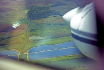 An aerial view showing vast stretches of green fields interspersed with a grid of blue solar panels. A small body of water is visible near the center of the fields. Part of an airplane's wing and engine are visible in the foreground, indicating the image is taken from an airplane.