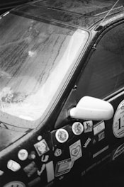 A close-up view of a car covered in a variety of stickers. The window is slightly fogged and has water droplets. The side mirror is visible, and the car's surface appears wet, indicating a rainy or damp environment.