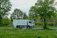 A white garbage truck is parked on a road adjacent to a lush, green park. Trees with vibrant green leaves provide a natural backdrop, and the ground is covered with grass and scattered white flowers.
