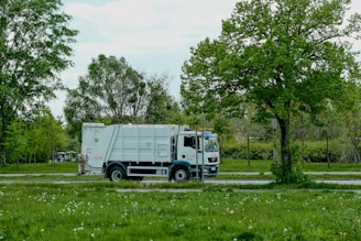 A white garbage truck is parked on a road adjacent to a lush, green park. Trees with vibrant green leaves provide a natural backdrop, and the ground is covered with grass and scattered white flowers.