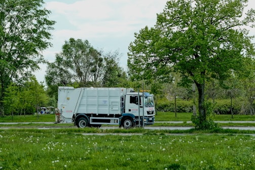 A white garbage truck is parked on a road adjacent to a lush, green park. Trees with vibrant green leaves provide a natural backdrop, and the ground is covered with grass and scattered white flowers.