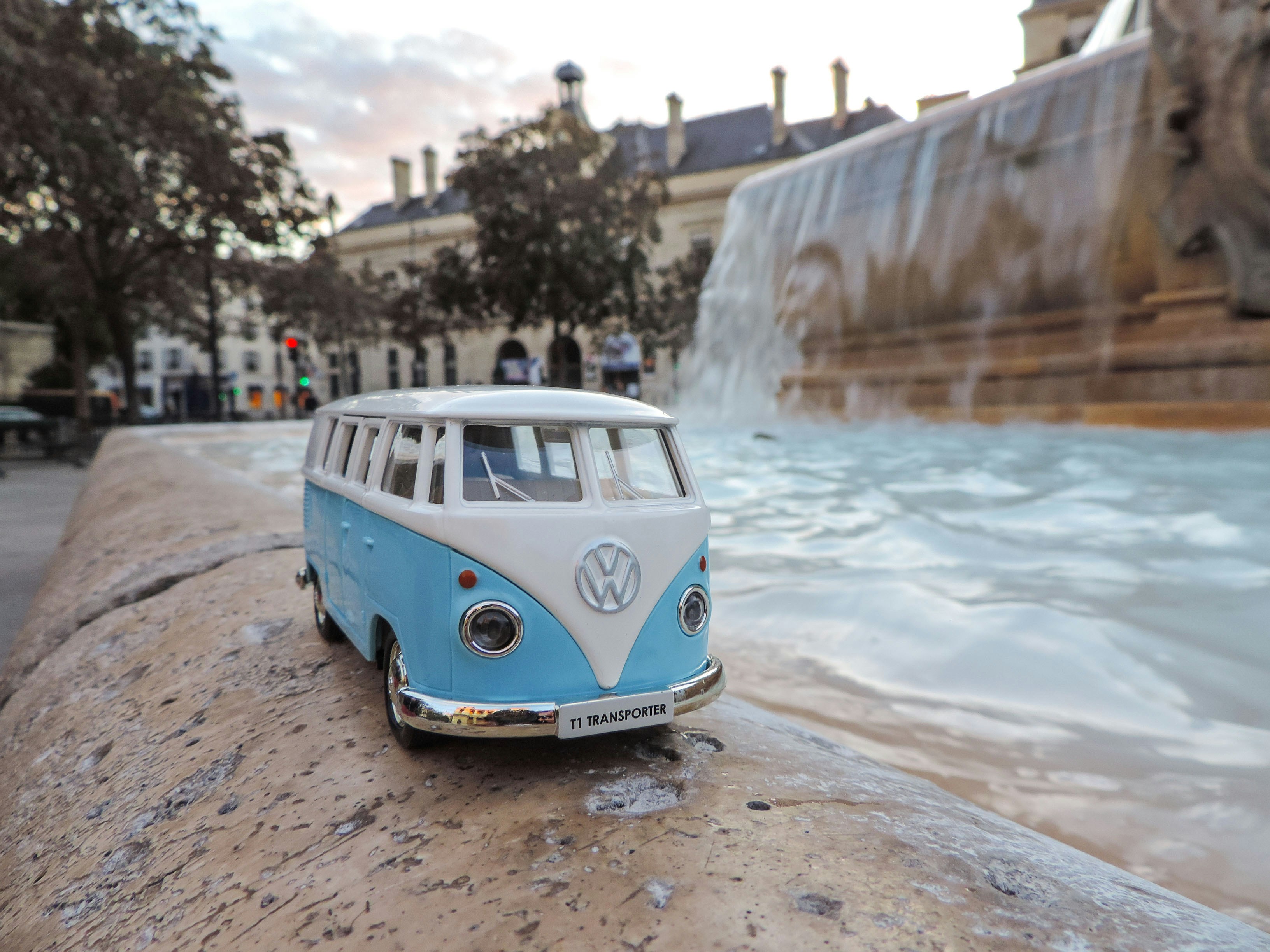 a car parked on a road by a body of water, Blue toy Volkswagen T1 with the Fontaine Saint-Sulpice at background. Sunny day outside.