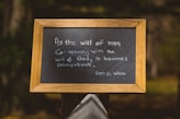 A framed blackboard with handwritten text that reads: 'As the will of man co-operates with the will of God, it becomes omnipotent. Ellen G. White.' The board has a wooden frame and is placed outdoors with a blurred natural background.