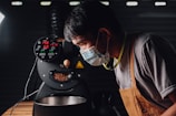 A person wearing a mask and an apron is closely observing a coffee roasting machine. The machine's display shows various numbers related to temperature and time, and coffee beans are visible inside. The environment suggests an industrial or professional setting with dim lighting.